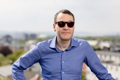 Mann med mørke solbriller, mørkt hår og blå skjorte. Han står ute.