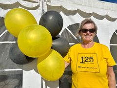 En smilende kvinne med solbriller og gul t-skjorte det står "125 år 1900-2025 Norges Blindeforbund" på, står ved siden av gule og svarte ballonger.