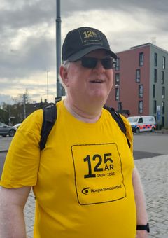 En mann med solbriller, sort caps og en gul t-skjorte det står "125 år 1900-2025 Norges Blindeforbund" på.