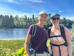 To smilende damer med caps og turtøy står ved en elv i et landlig landskap.