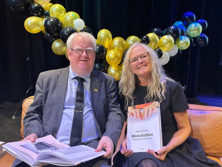 En smilende mann og kvinne, begge med briller, sitter ved siden av hverandre og holder i hver sin store bok. Bak dem henget det mange gule, og sorte ballonger.
