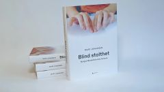 En stor bok med bilde av et par hender som leser punktskrift og tittelen "Blind stolthet - Norges Blindeforbunds historie" står foran en stabel med tre eksemplarer av samme bok.