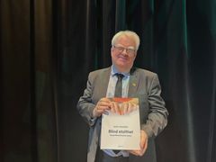 En smilende mann i dress og med briller holder en stor bok som det står "Blind stolthet" på.