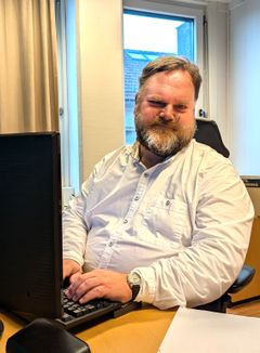 En mann med skjegg, bart og i hvit skjorte sitter ved et skrivebord med en datamaskin.