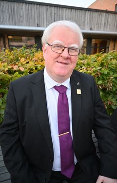 En mann med hvitt hår, briller, dress og lilla slips.
