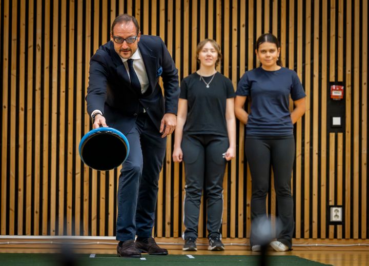 Kronprins Haakon står sammen med to jenter i en gymsal og svinger på en curlingsten.