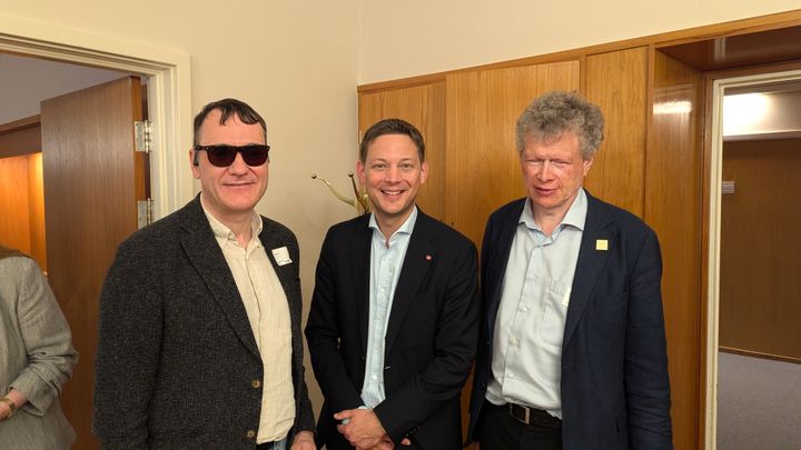 Tre menn står inne. De smiler. Han til venstre har mørke briller