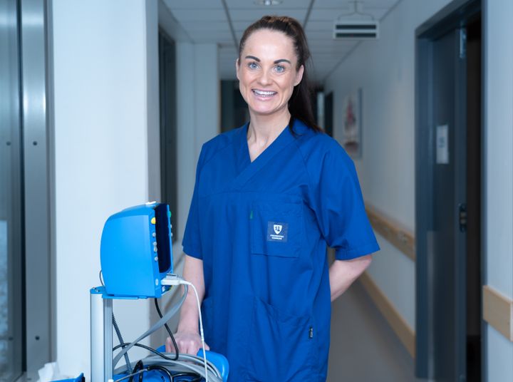 Person i blå arbeidsuniform står i en korridor i en helseinstitusjon ved siden av medisinsk utstyr.