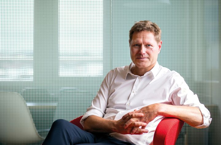 Person%20i%20hvit%20skjorte%20sitter%20tilbakelent%20p%E5%20en%20r%F8d%20stol%2C%20i%20et%20kontorlokale%20med%20glassvegg%20i%20bakgrunnen.