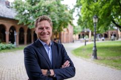 Mann i dress som smiler. Litt bakoverlent med armene samlet foran kroppen. Utendørs på en brosteinsbelagt gangvei med parkmiljø i bakgrunnen.