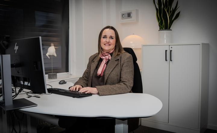 Kvinne%20som%20sitter%20ved%20en%20pult%2C%20som%20ser%20i%20kamera%20mens%20hun%20skriver%20p%E5%20tastaturet.