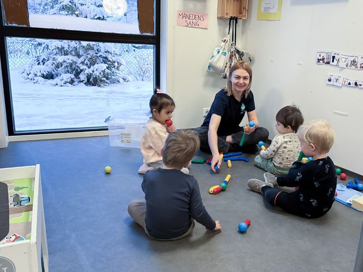 En kvinne sitter på gulvet og leker med tre små barn i en barnehage. Snø kan sees gjennom vinduet i bakgrunnen.