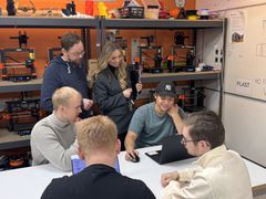 Personer samlet rundt et bord i et verkstedmiljø med 3D-printere i bakgrunnen. Flere ser på en bærbar PC mens én person bruker mus. Hyller med teknisk utstyr står langs veggene.