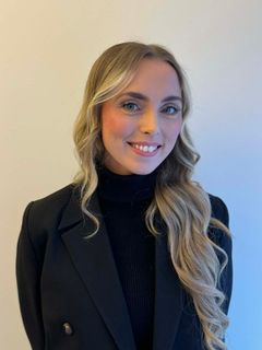 Portrettfoto av Marianne Bjerkmansett fra brystet og opp, mot lys bakgrunn. Personen ser mot kamera og har på seg mørk blazer og mørk høyhalset genser.