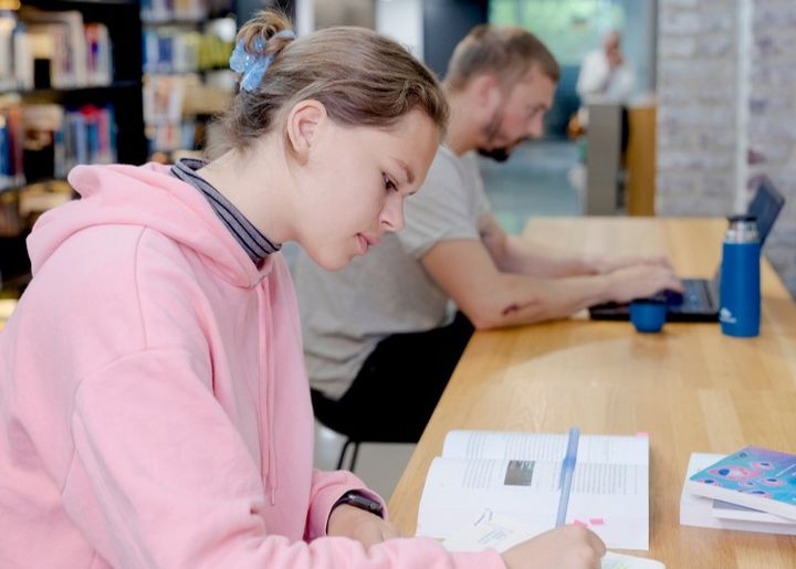 Ung kvinne og ung mann jobber med egenstudier på campus og bruker hhv. lærebøker og laptop