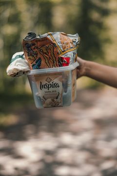 En person holder en plastboks fylt med søppel på en grusvei ute i naturen.