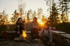 Tre personer sitter rundt et bål i skogen ved solnedgang.