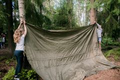 To personer setter opp en grønn presenning mellom trær i skogen.