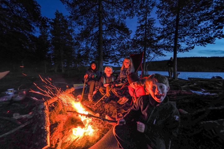 En gjeng med barn sitter samlet rundt varmen fra bålet i kveldsmørket.