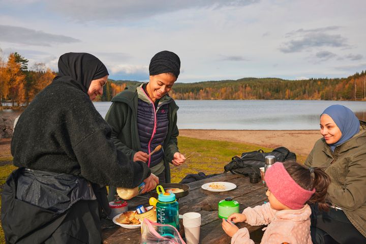 Tre kvinner og ei jente er samlet rundt et bord og holder på med matlaging, ut ved et vann.