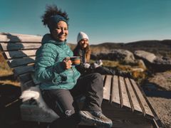 To damer sitter på en benk i naturen og nyter hver sin kopp med varm drikke og smiler.