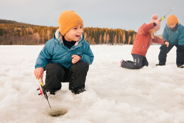 En liten gutt sitter på huk og isfisker med et smil om munnen