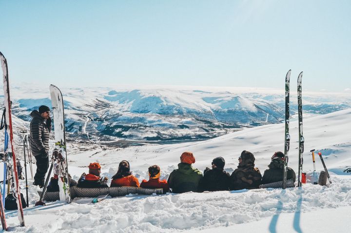 Bilde av en rekke folk som sitter mellom to oppsatte par med ski og skuer utover et vinterkledd fjellandskap