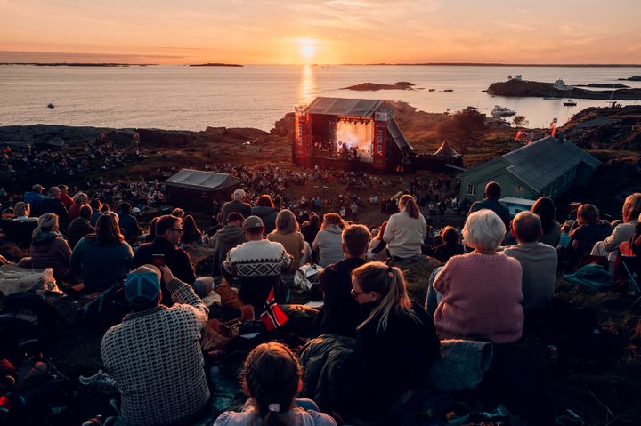 Et stort publium sitter vendt mot en utendørs scene på Fløløy i storslått solnedgang.