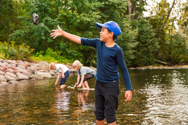En gutt står til knes i vannkanten og kaster en stein.