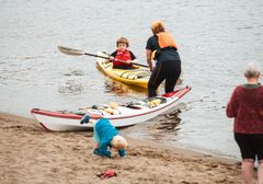 En gutt padler i kajakk på en strand, mens en voksen hjelper til. En baby krabber på sanden.