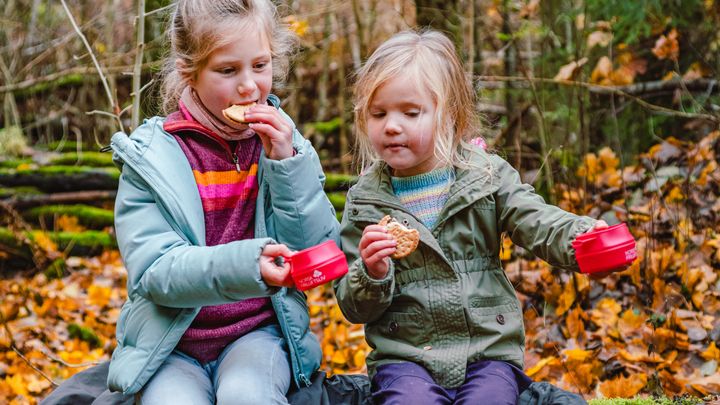 To jenter med hver sin kjeks i hånda og røde turkopper, sitter på en benk. I bakgrunnen er det skog og løv i høstfarger.